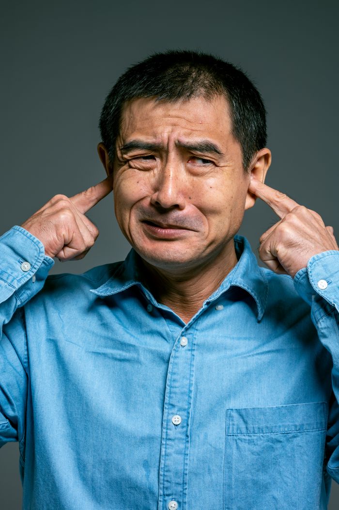 Asian man in blue denim shirt expressing frustration by covering his ears in a close-up portrait.