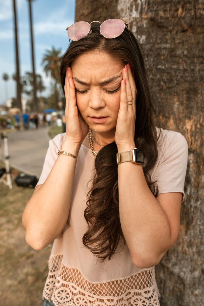 Young woman with long hair stands by a tree with a headache, emphasizing stress relief outdoors.