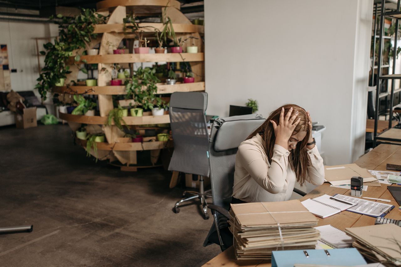 Services A stressed woman sits overwhelmed at her desk, surrounded by paperwork in a modern office setting.